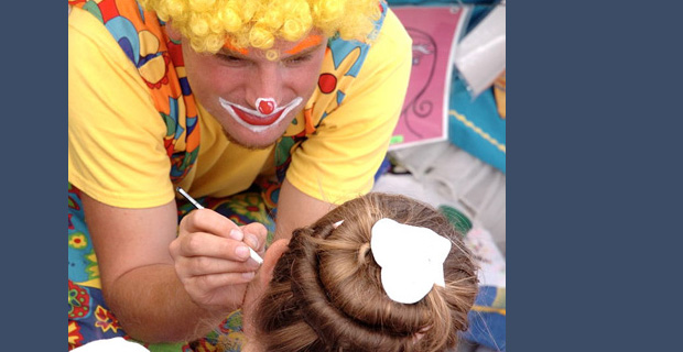 A little girl getting her face painted by a clown.